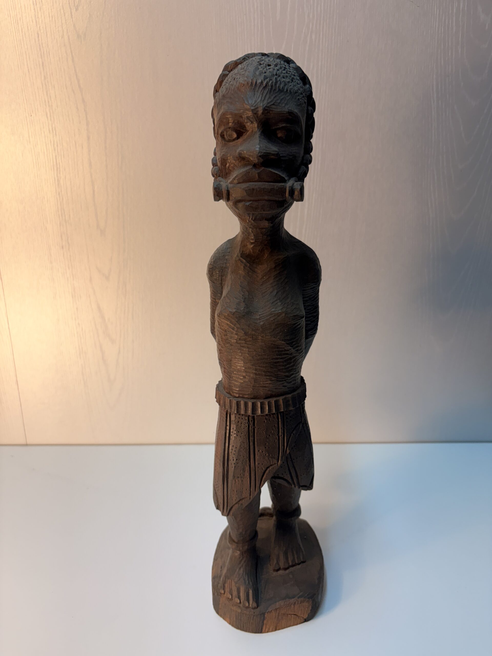 full view of an African wooden figure with restrained hands and a covered mouth